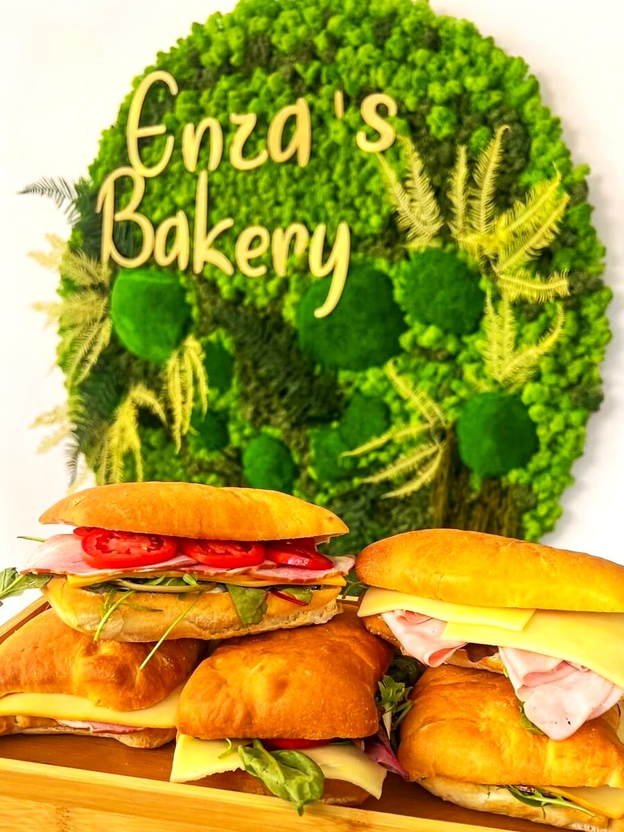 Panini pe tocător de lemn, în fața peretelui de mușchi verde cu sigla Enza's Bakery